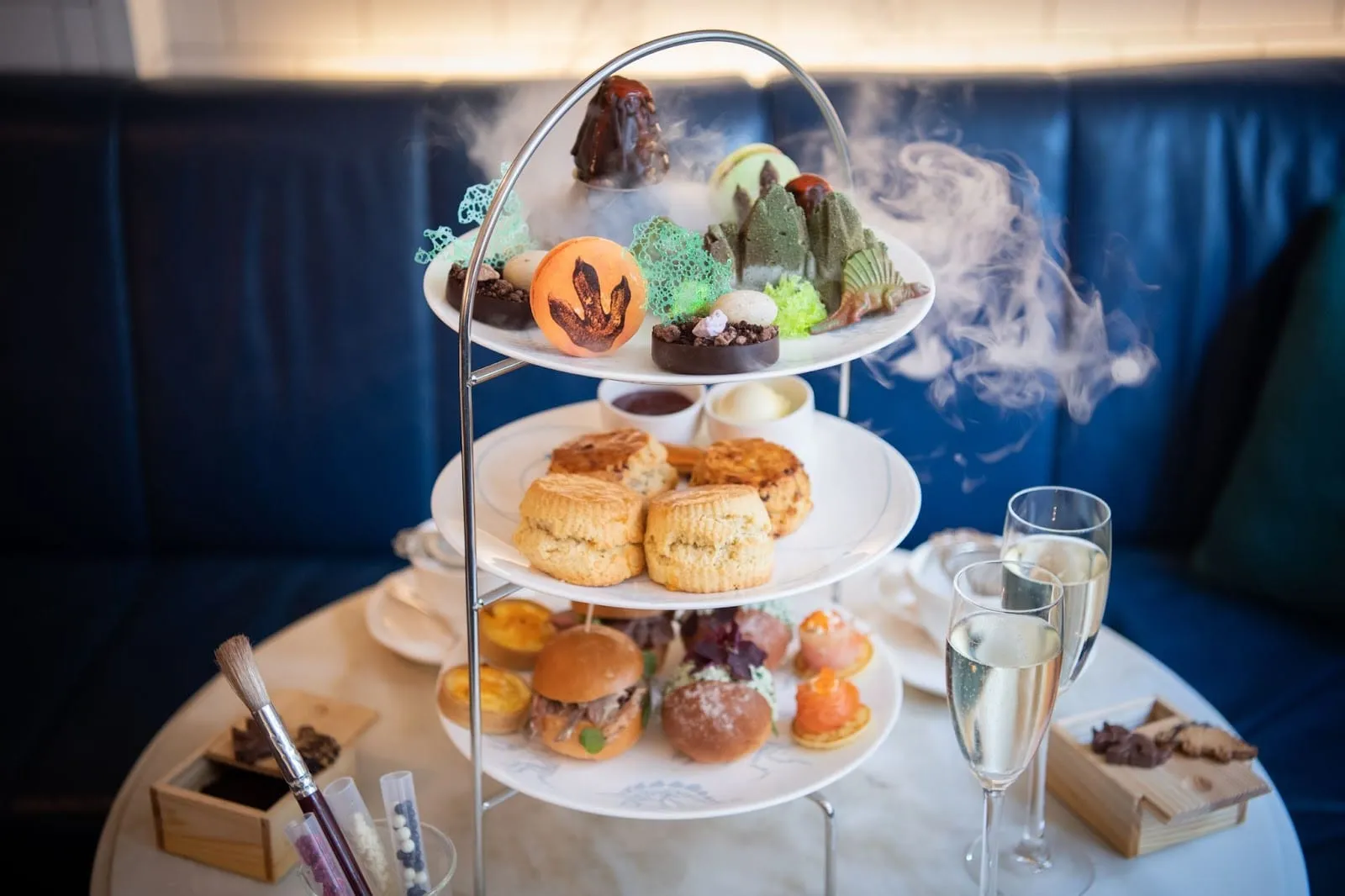 A three-tiered Jurassic afternoon tea stand wreathed in dry ice at The Ampersand, featuring a T-rex footprint macaron, chocolate dinosaur volcano, and fossil excavation kit