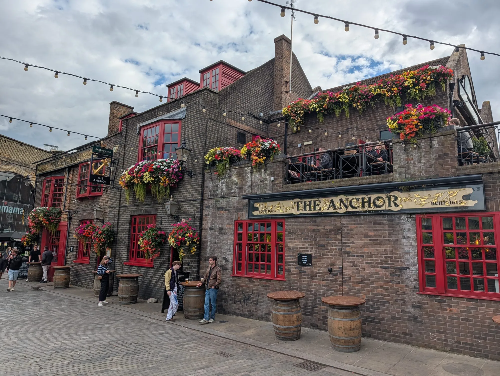 The Anchor Bankside, a brick riverside pub hung with flower baskets, with barrel tables outside