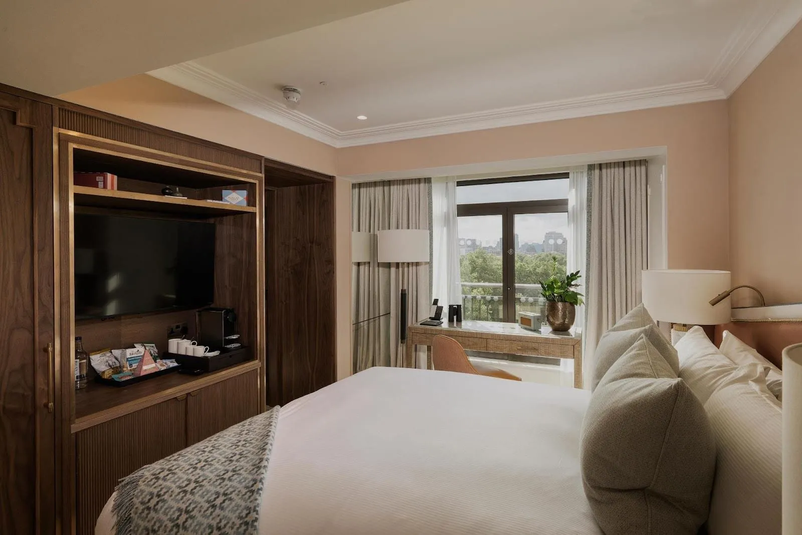An Athenaeum bedroom with a walnut cabinet holding a television and tea tray, a desk by the window, and a balcony door looking over the trees of Green Park