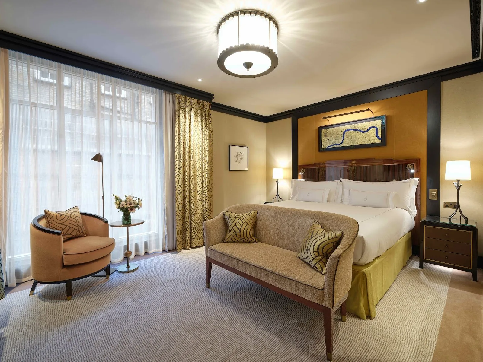 A bedroom at The Beaumont Mayfair with a mustard velvet headboard, art deco lighting and a framed map of the Thames above the bed