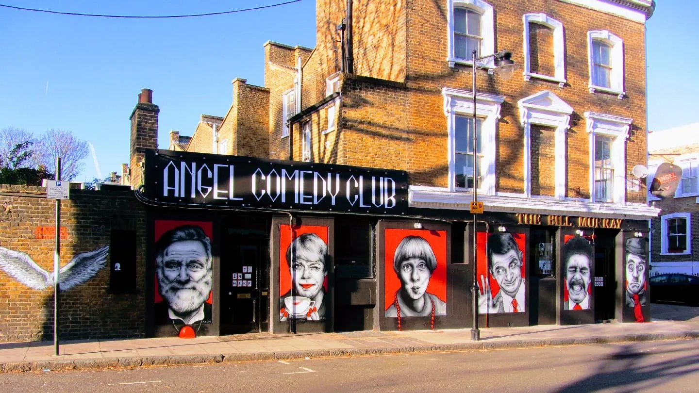 The corner exterior of The Bill Murray pub, with large painted portraits of comedians on red panels along the ground floor and the pub's name in gold lettering