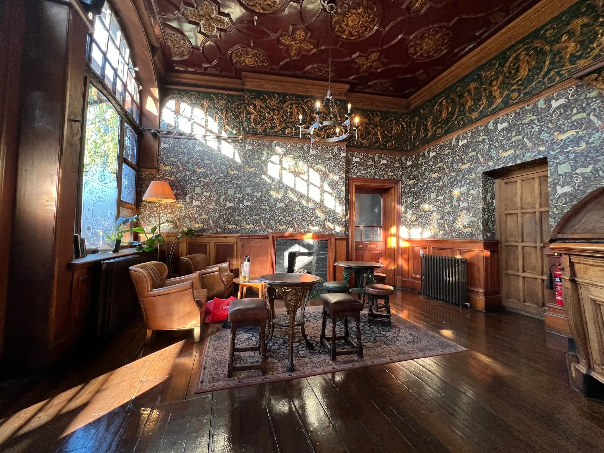 A room at The Black Lion in Kilburn with a deep red patterned ceiling, a decorative frieze and cornice, patterned wallpaper, armchairs and sunlight on a wooden floor