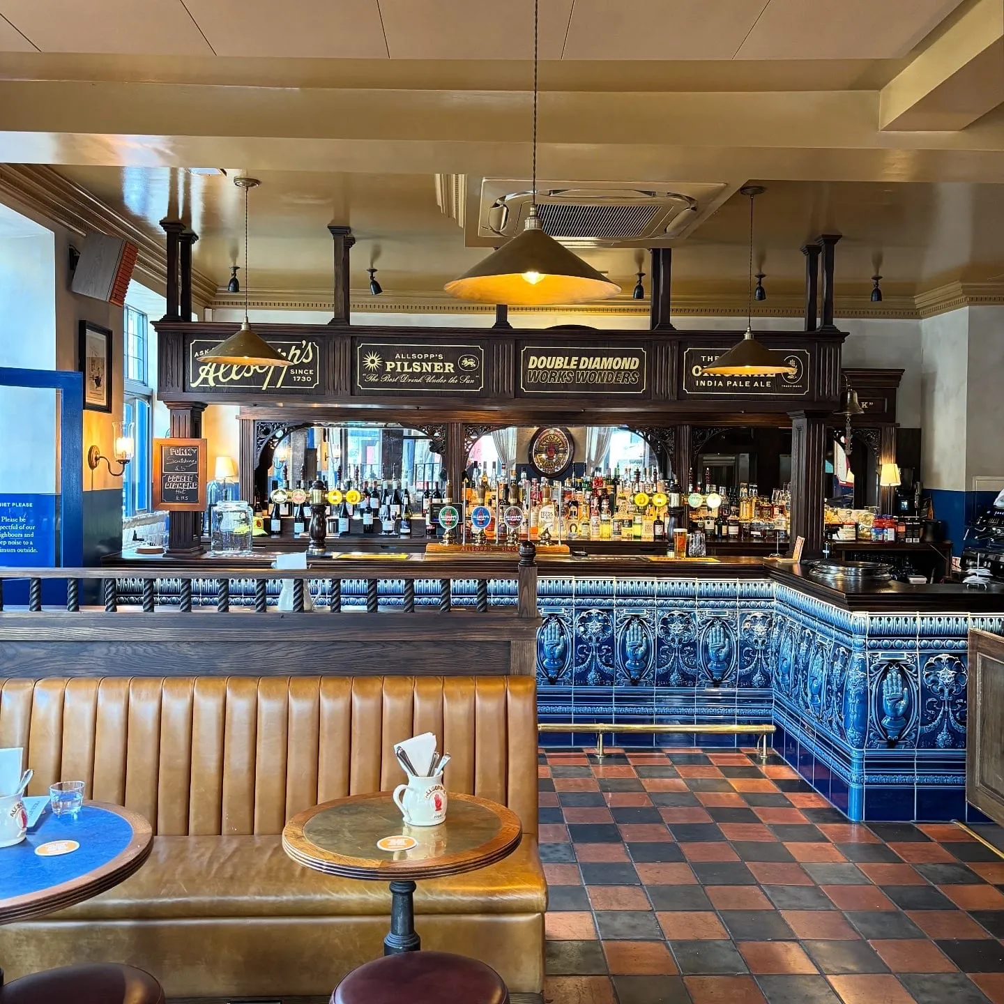 A dark wood bar back with gold Allsopp's brewery lettering, above a blue ceramic bar front with a raised hand emblem, at The Blue Stoops
