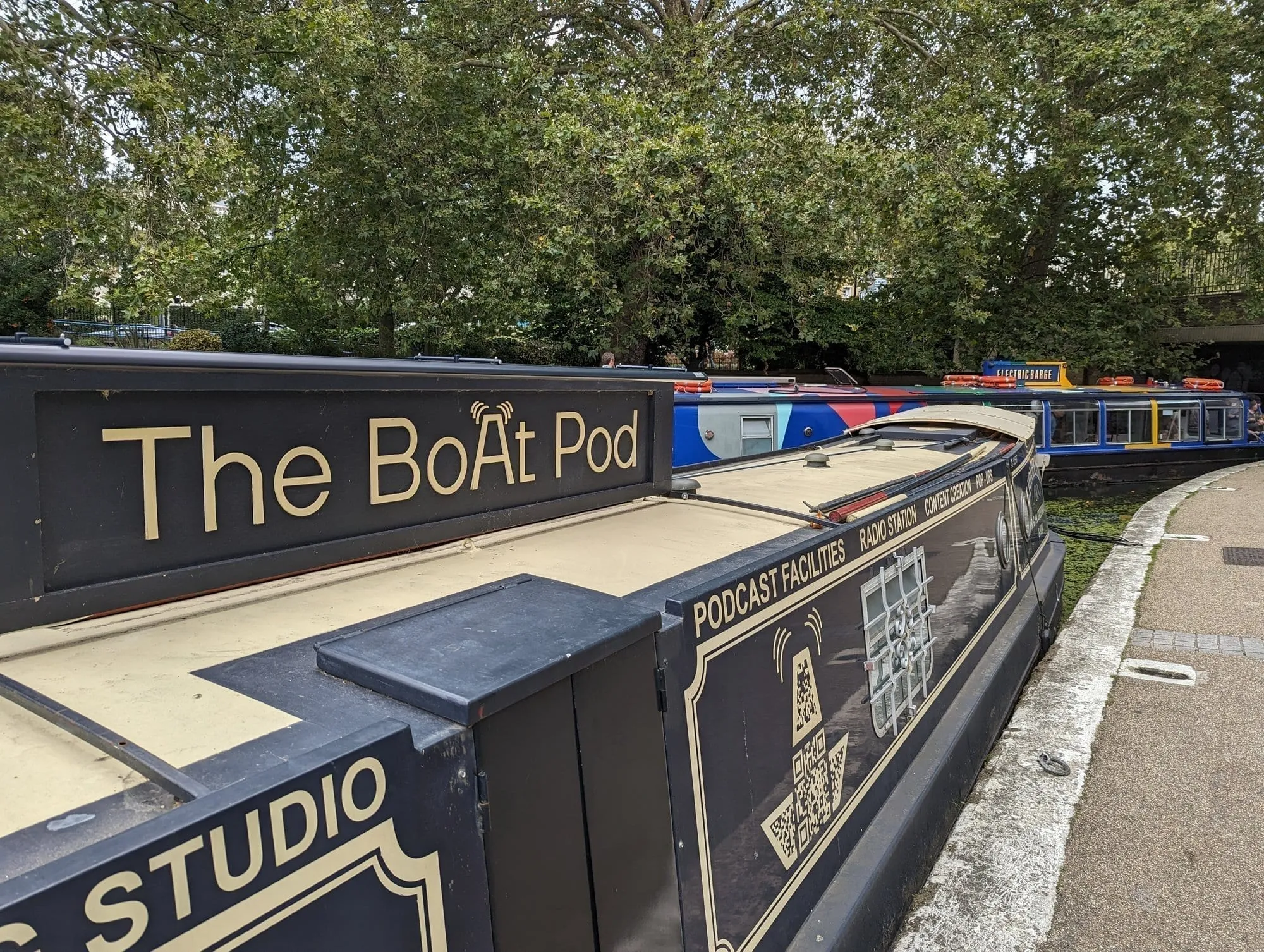 The gold lettered "The BoAt Pod" signage on the roof of a black narrowboat moored at Little Venice, with "Podcast Facilities" and "Studio" painted on the side