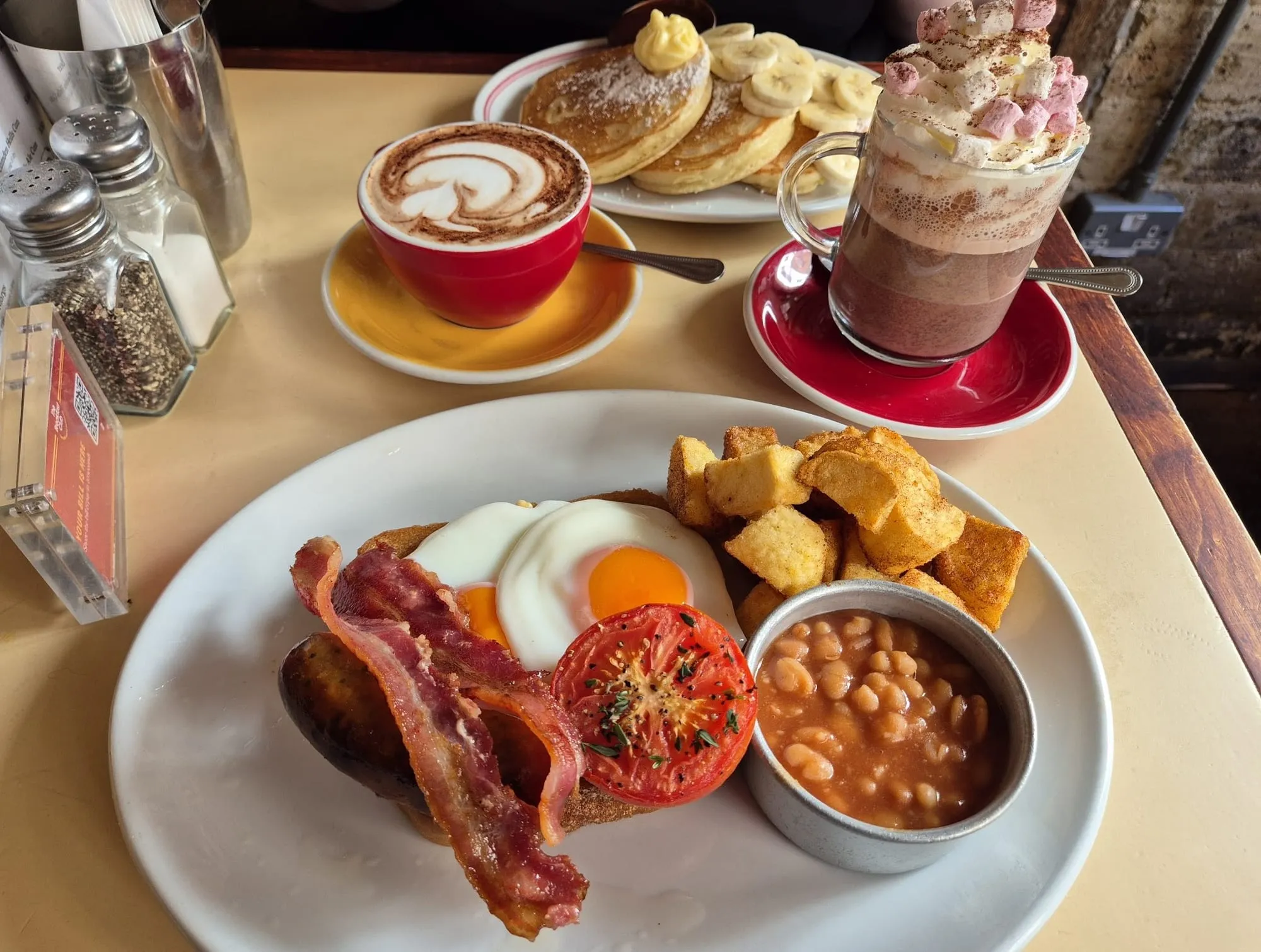 A full breakfast with poached eggs, bacon and beans next to a stack of pancakes with banana and a hot chocolate at The Breakfast Club
