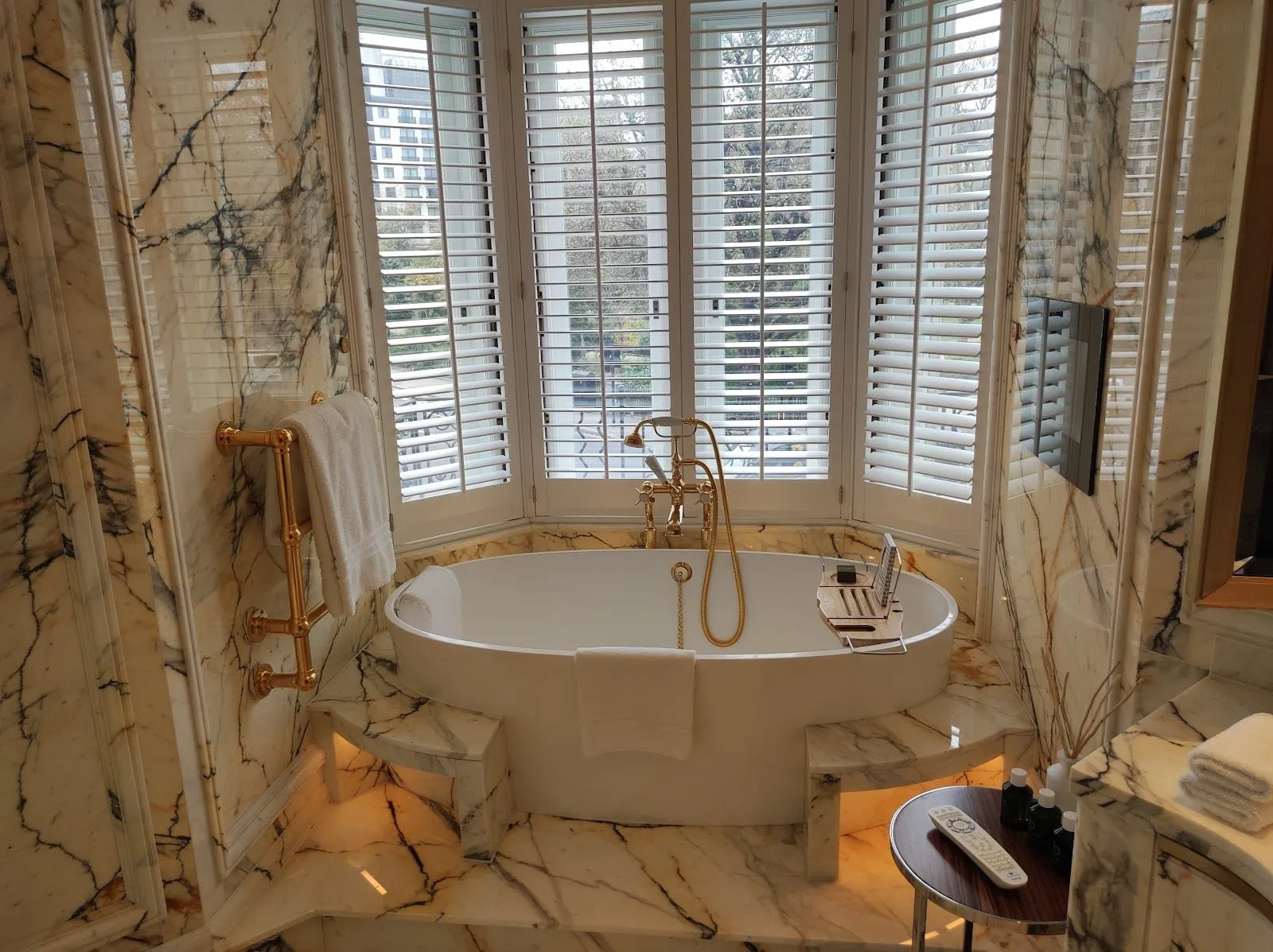 A suite bathroom at The Cadogan: a freestanding bath on a veined marble plinth in a shuttered bay window over trees, with gold taps and a gold towel rail
