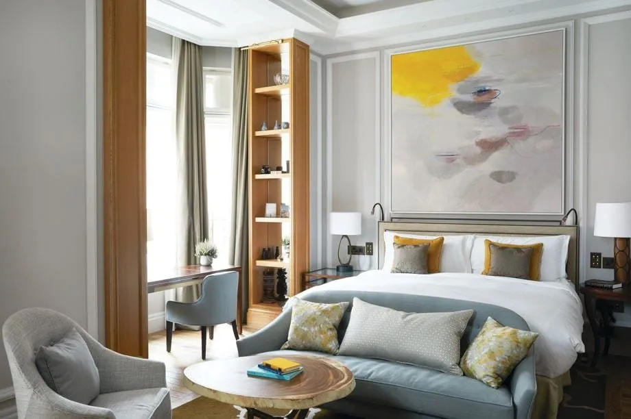A room at The Cadogan with pale grey panelled walls, a large abstract painting in grey and yellow above the bed, a pale blue sofa at the foot of the bed, a desk in the bay window and oak shelves