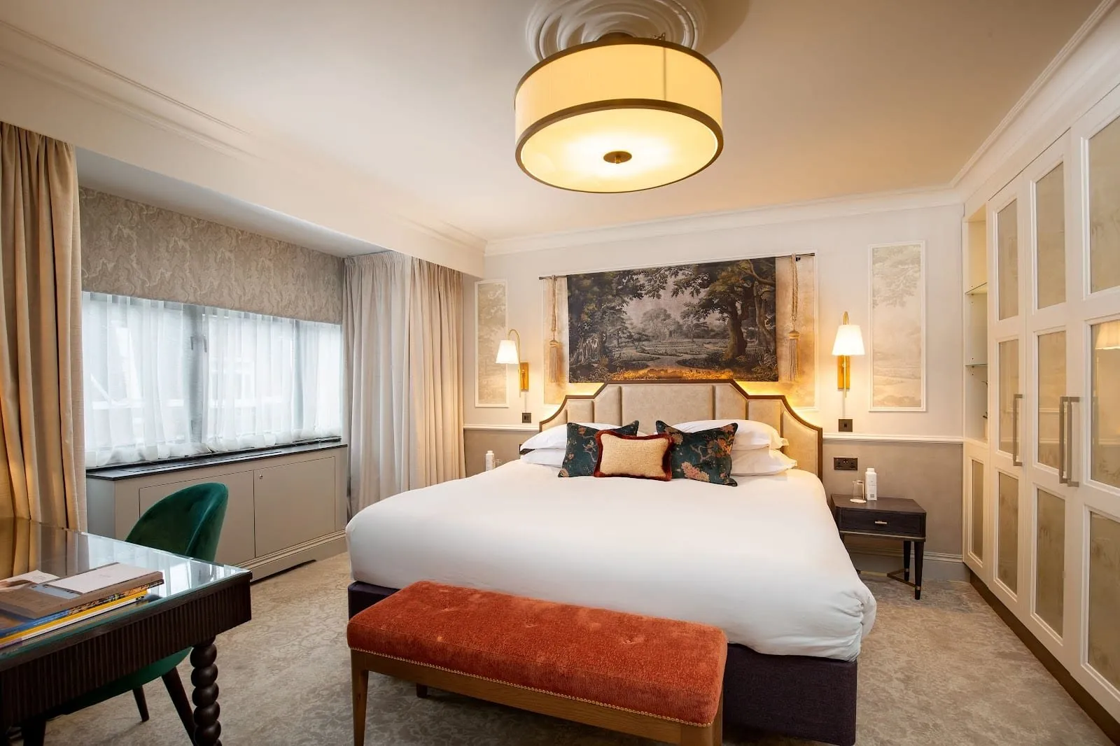 The bedroom of a Junior Suite at The Capital: a king bed with a cream upholstered headboard beneath a tapestry of trees, brass wall lights, an orange velvet bench, a green velvet desk chair and a wide window with sheer curtains