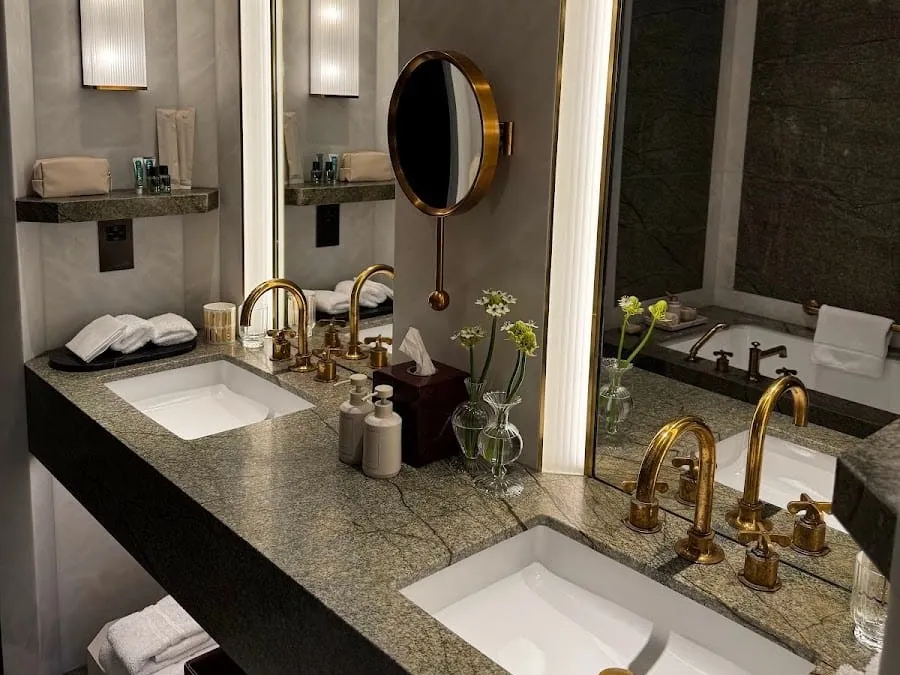 A Chancery Rosewood bathroom with twin basins set into a green-veined stone counter, brass taps, lit wall mirrors and a bath reflected behind