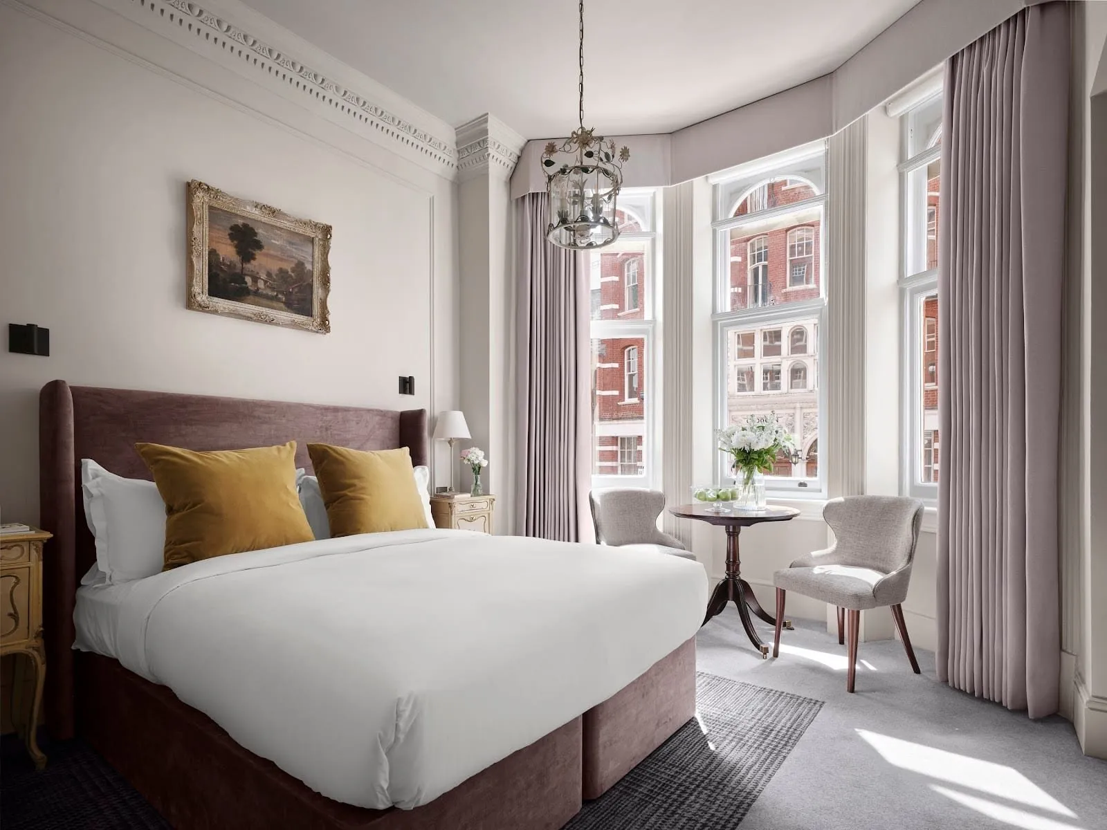 A Chelsea Double at The Chelsea Townhouse: a bed with a plum velvet headboard and mustard cushions, a gilt-framed landscape painting, a lantern chandelier and a bay window with a small table for two, facing red-brick buildings