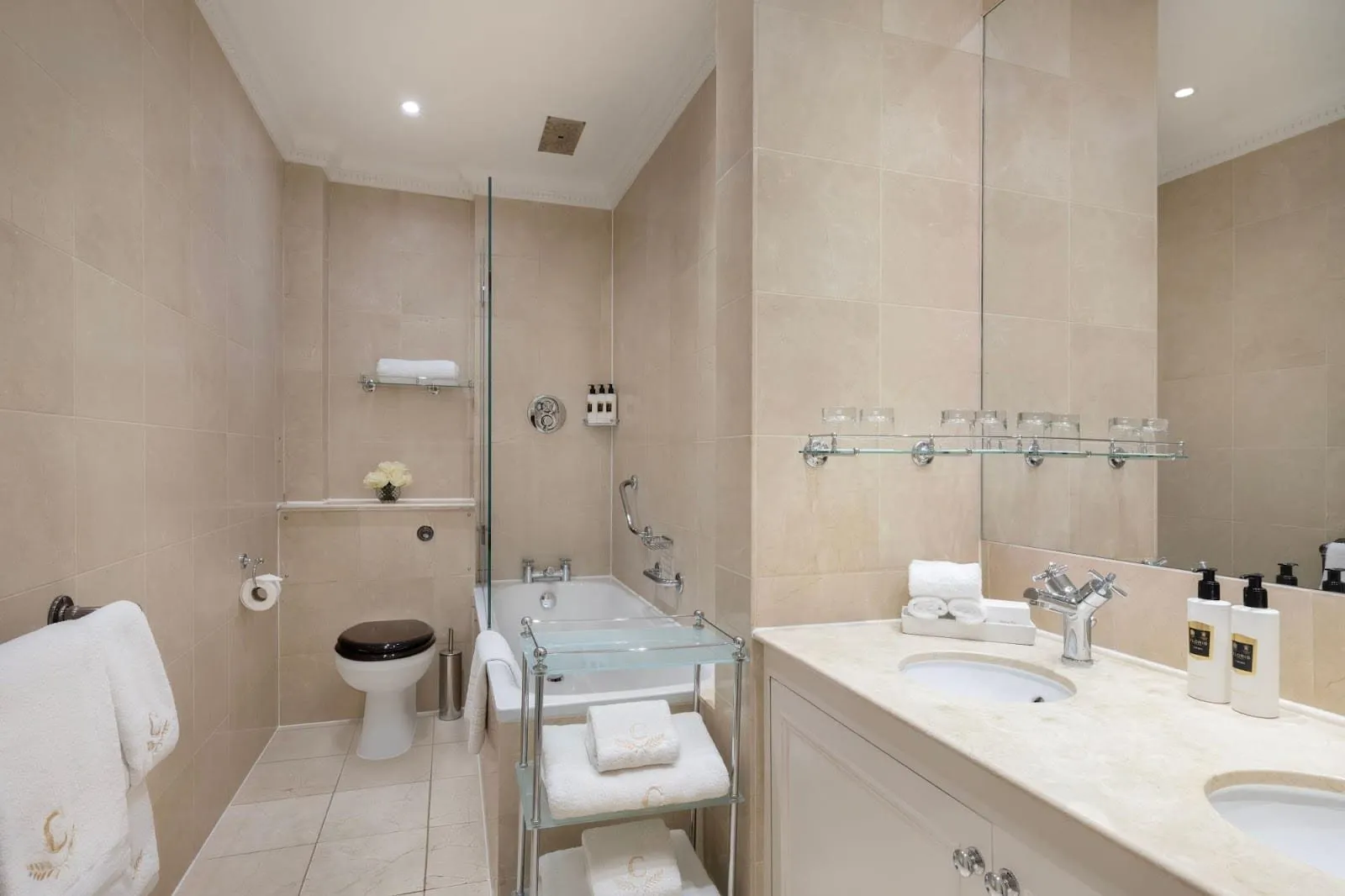 A Chesterfield bathroom in cream marble with a bath under a glass screen and ceiling shower, a WC, twin basins and towels embroidered with a gold C