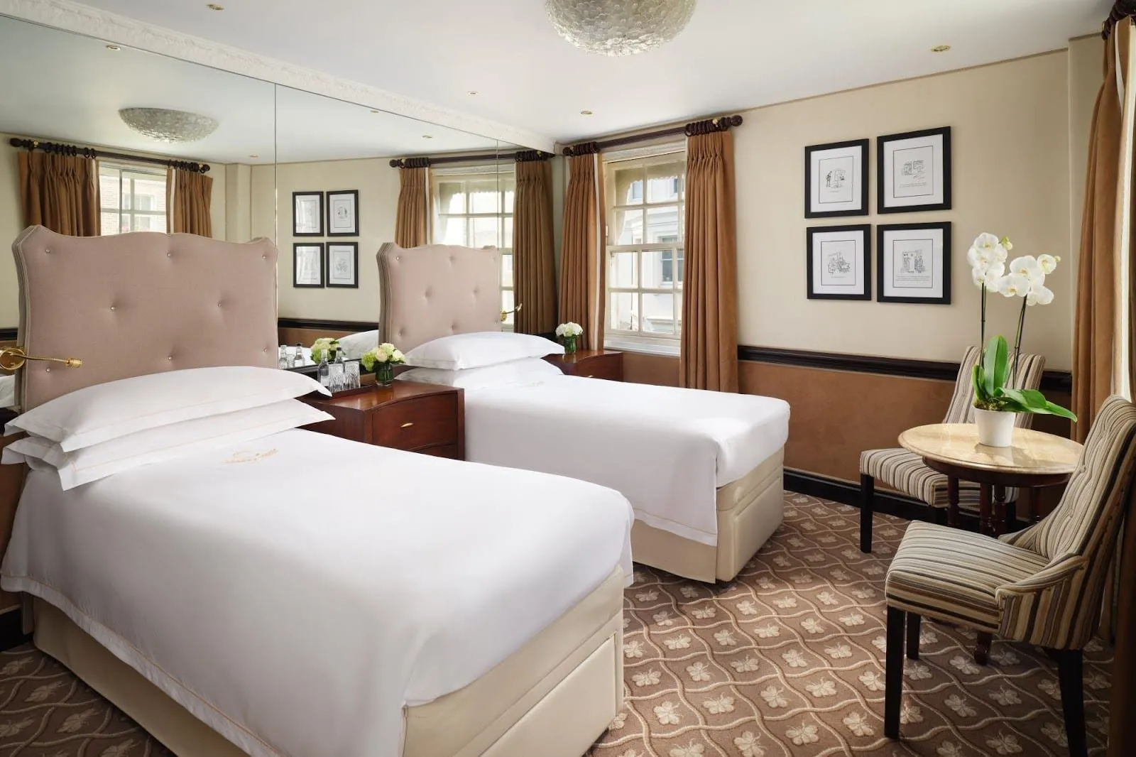 A twin room at The Chesterfield with two single beds under buttoned beige headboards, a mirrored wall, framed cartoons, striped chairs at a marble-topped table and two sash windows