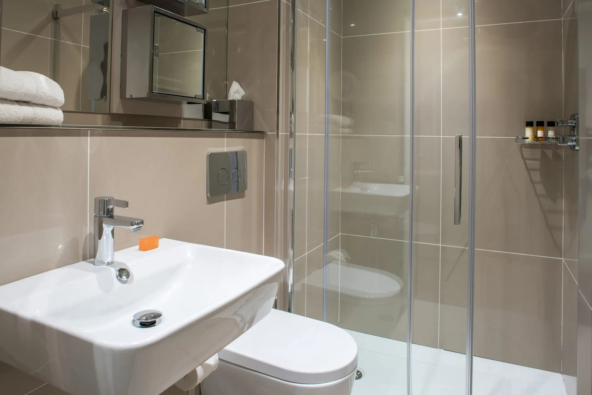 The bathroom at The Chronicle, with beige tile, a glass-enclosed shower and a mirrored cabinet
