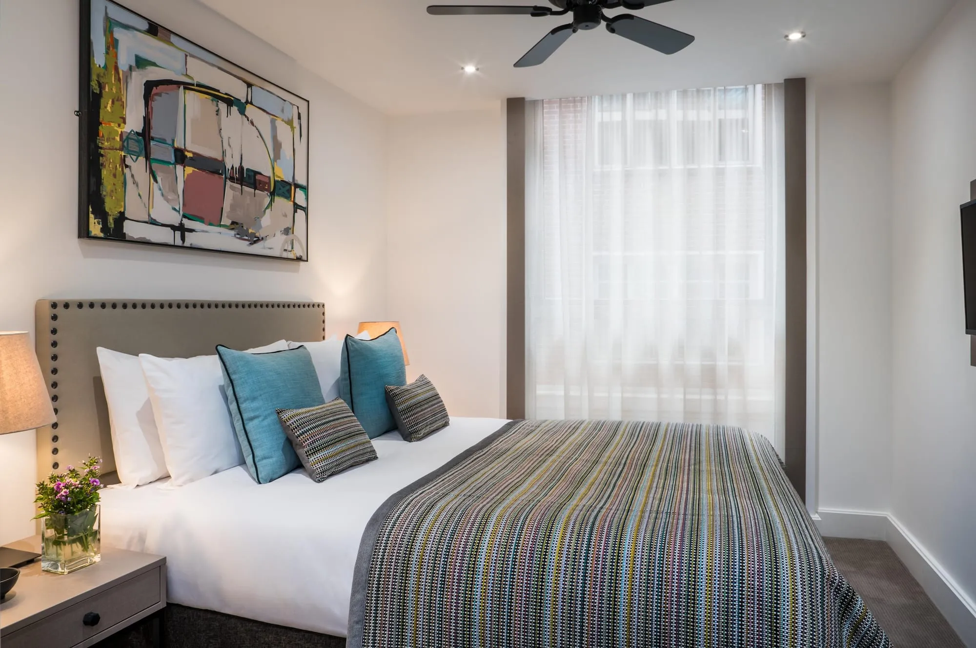 A bedroom at The Chronicle, with a grey buttoned headboard, teal cushions and a striped bedspread beneath a ceiling fan