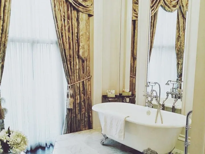 A white claw-foot roll-top bath on a marble floor at The Clermont, beside tall windows hung with gold damask curtains