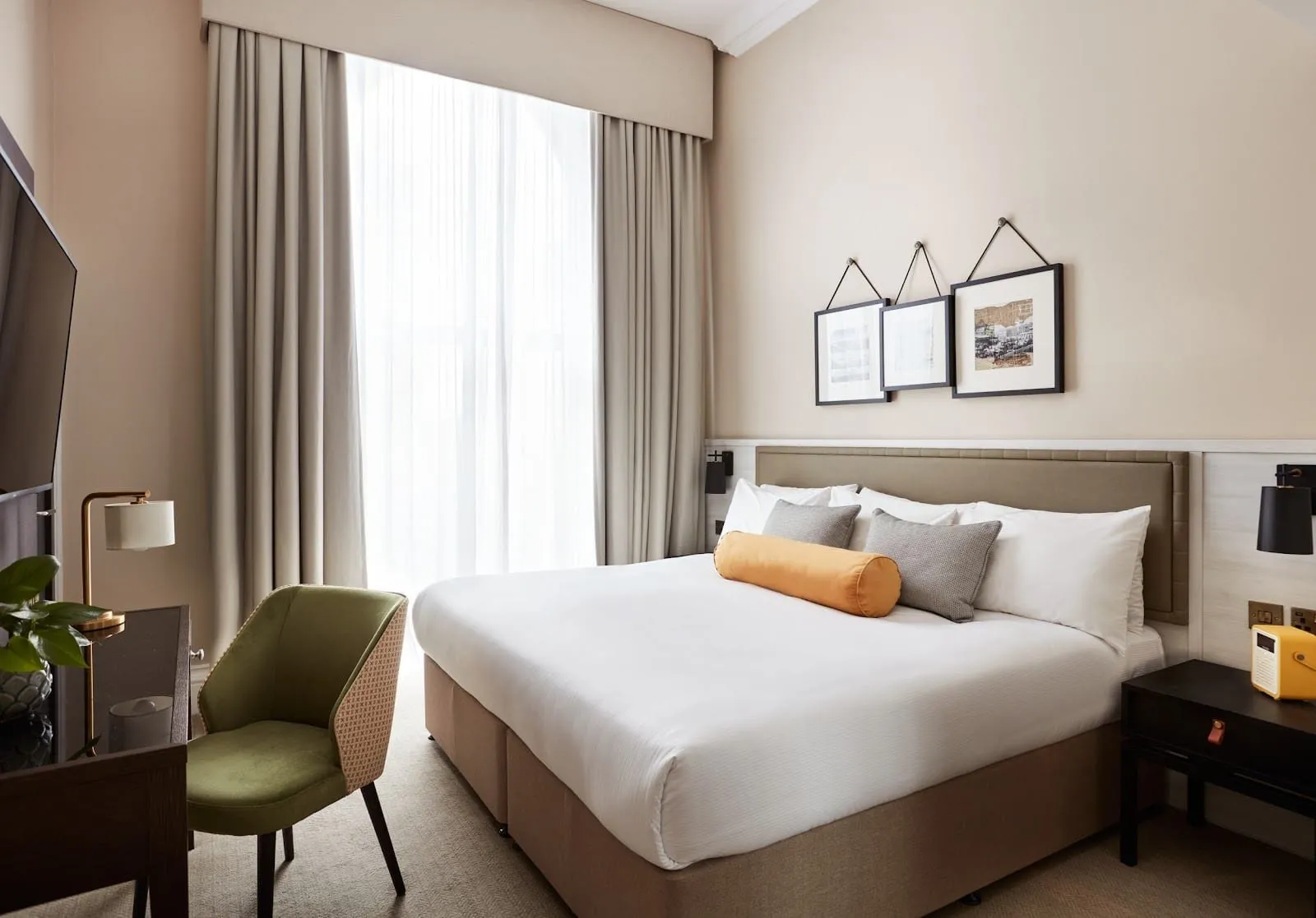 A double room at The Clermont London, Victoria, with a floor-to-ceiling window behind sheer curtains, a large double bed with a mustard bolster cushion, three framed prints hung on cords and a green velvet desk chair