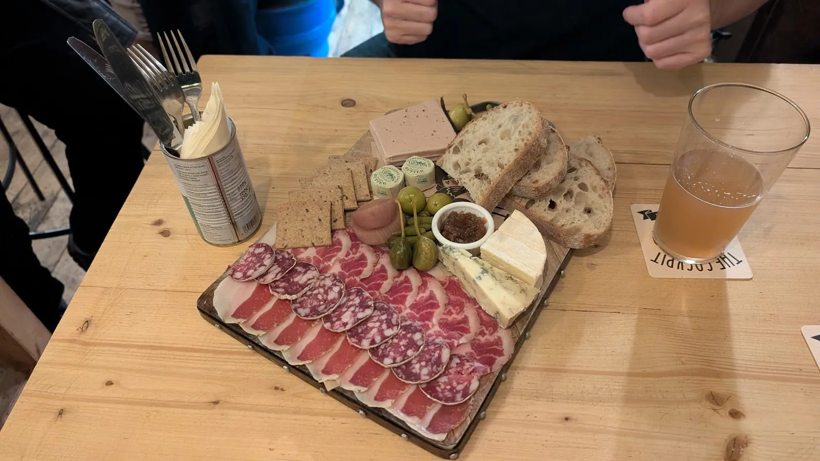A cheese and charcuterie board at The Cockpit, with cured meats, olives, bread and a wedge of blue cheese, next to a pint on a branded coaster