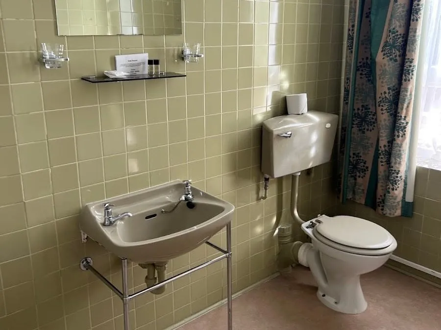 A 1970s bathroom at The Columbia with olive-green wall tiles, an avocado basin on a chrome washstand, a toilet with a separate cistern and a patterned curtain at the window