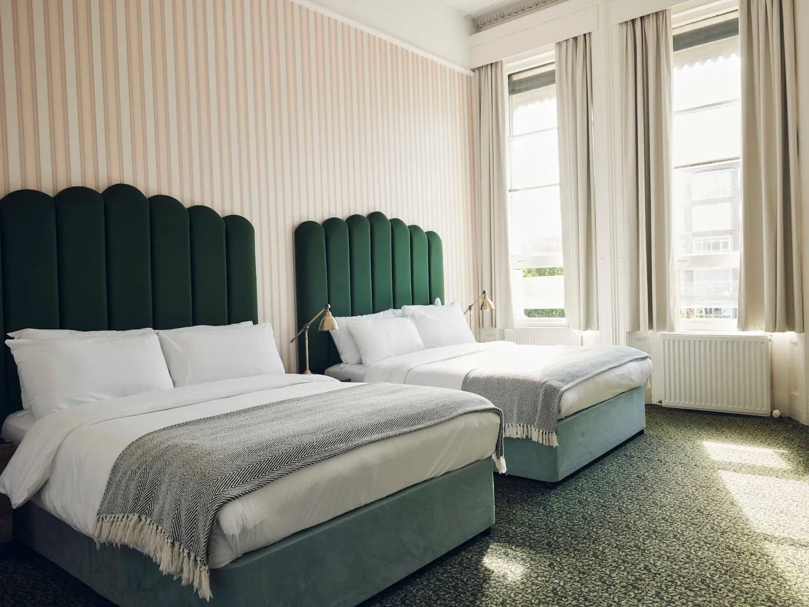 A family room at The Columbia with two double beds under tall scalloped green velvet headboards, striped wallpaper, a green patterned carpet and two tall sash windows