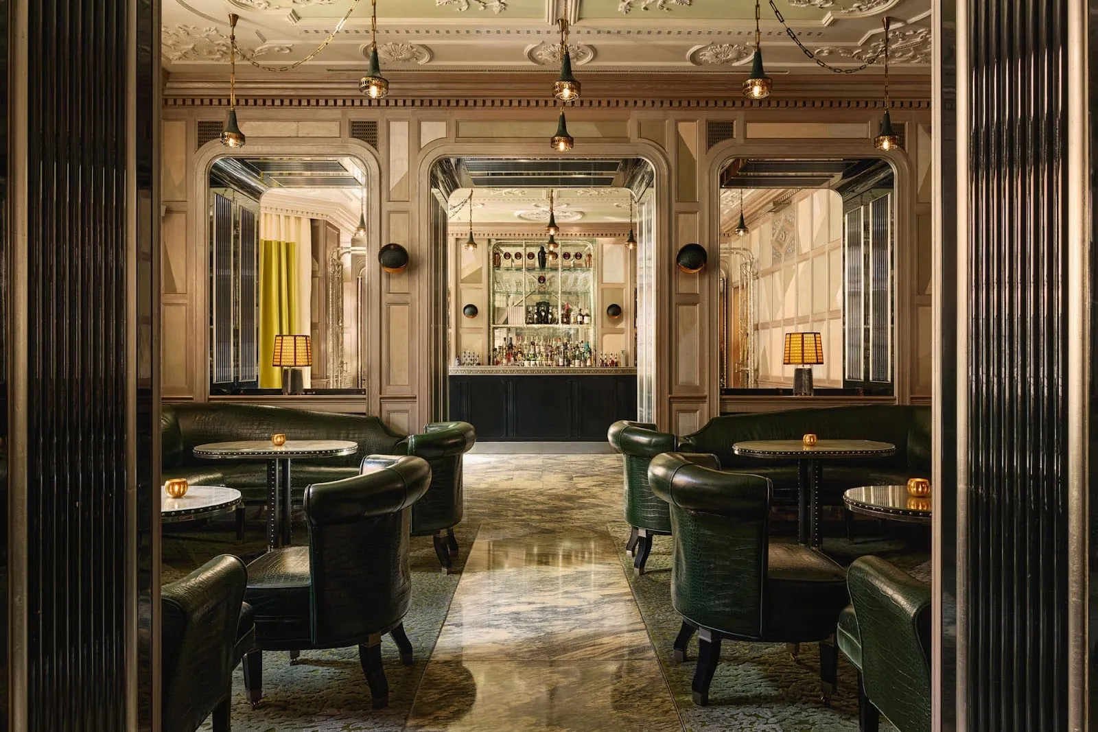 Green leather armchairs around small round tables in the Connaught Bar, with a lit back bar of bottles at the far end under a moulded plaster ceiling