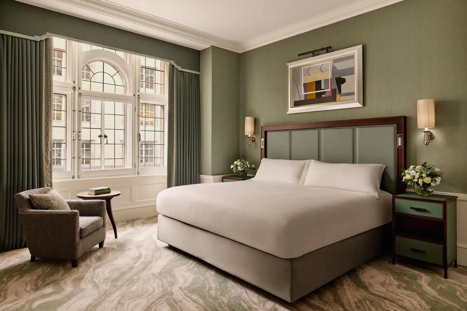 A Connaught bedroom in sage green with a panelled green leather headboard, an abstract painting above the bed, a grey armchair by a tall arched leaded window and a swirling green carpet