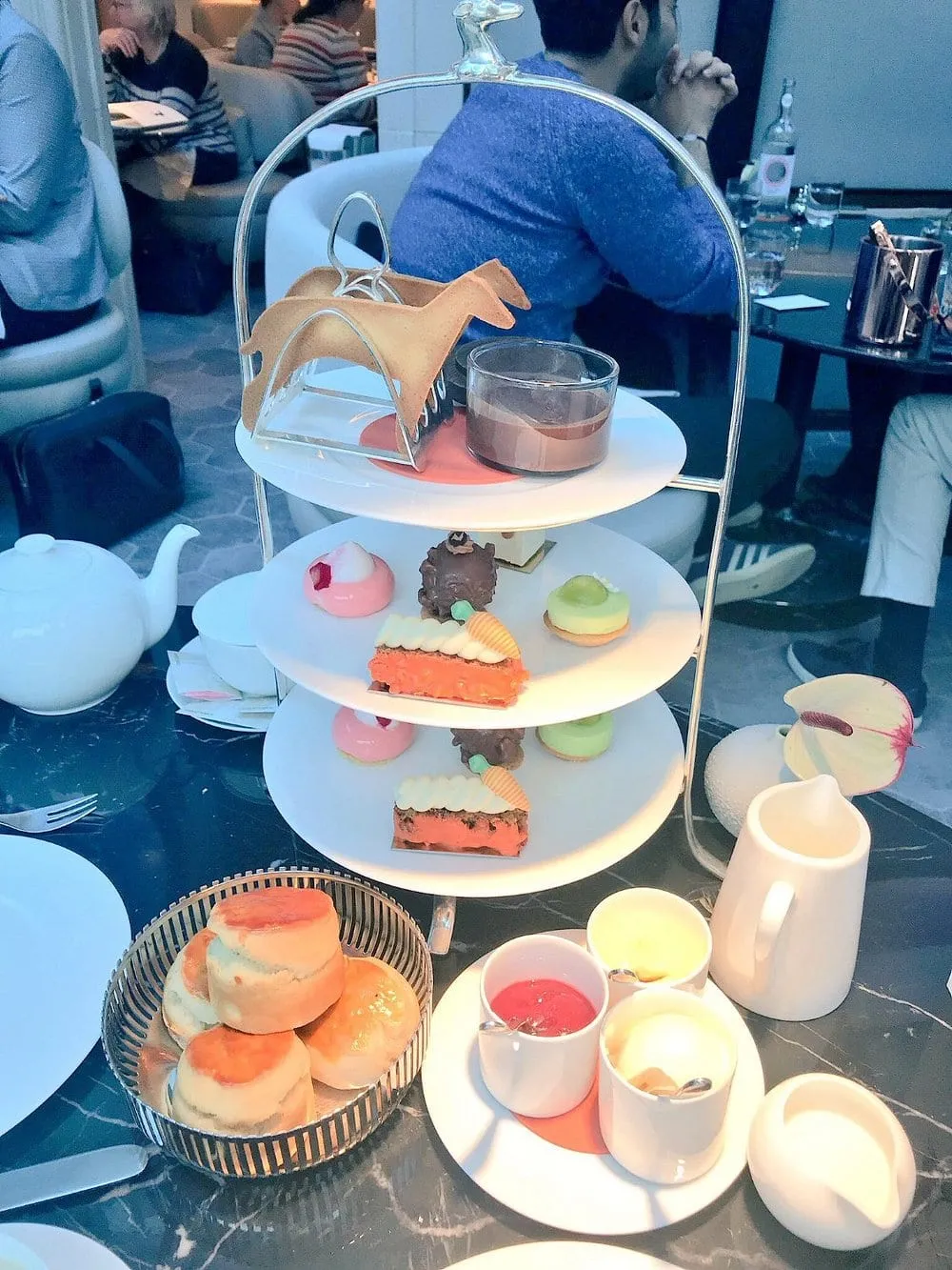 A tiered stand of pastries beside a silver basket of scones and pots of jam and cream on a dark marble table, with a horse-shaped biscuit on the top tier