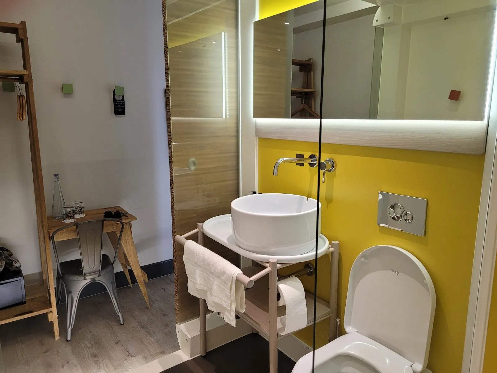 The ensuite bathroom of a room at The Corner London City, with a yellow accent wall, a vessel basin on a light wood stand and a walk-in shower