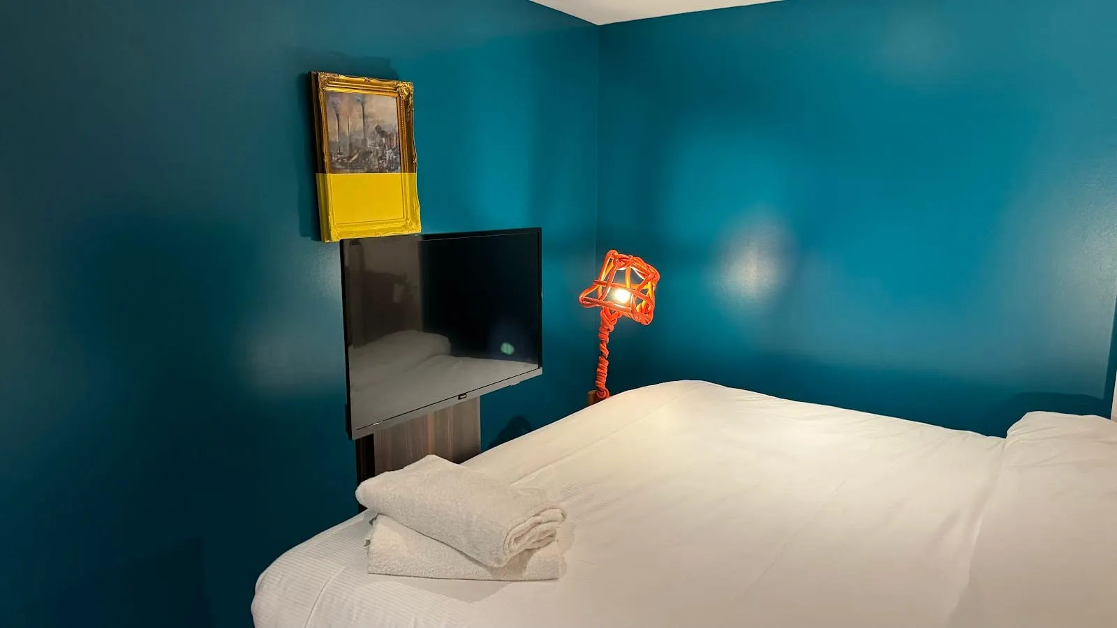 A room at The Corner London City with a teal-painted wall, a wall-mounted TV and a sculptural orange lamp