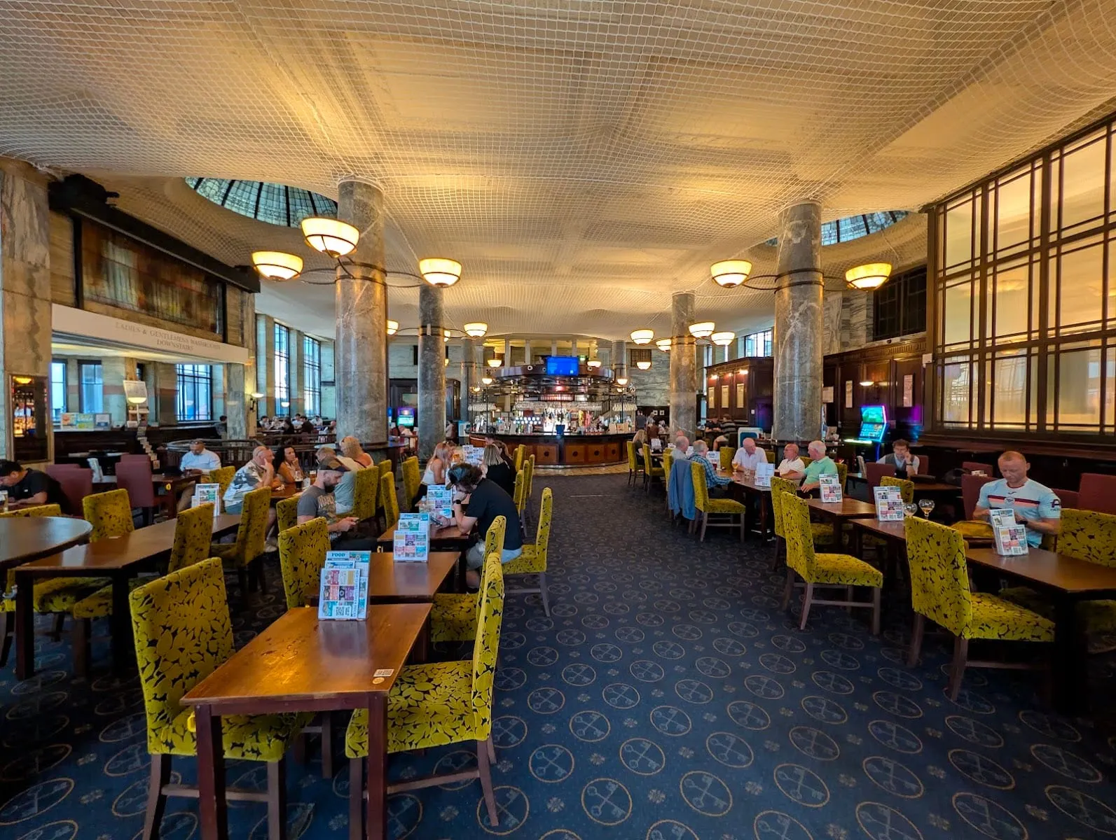 The banking hall interior of The Crosse Keys, with marble columns, hanging lamps and a stained-glass domed skylight above the bar