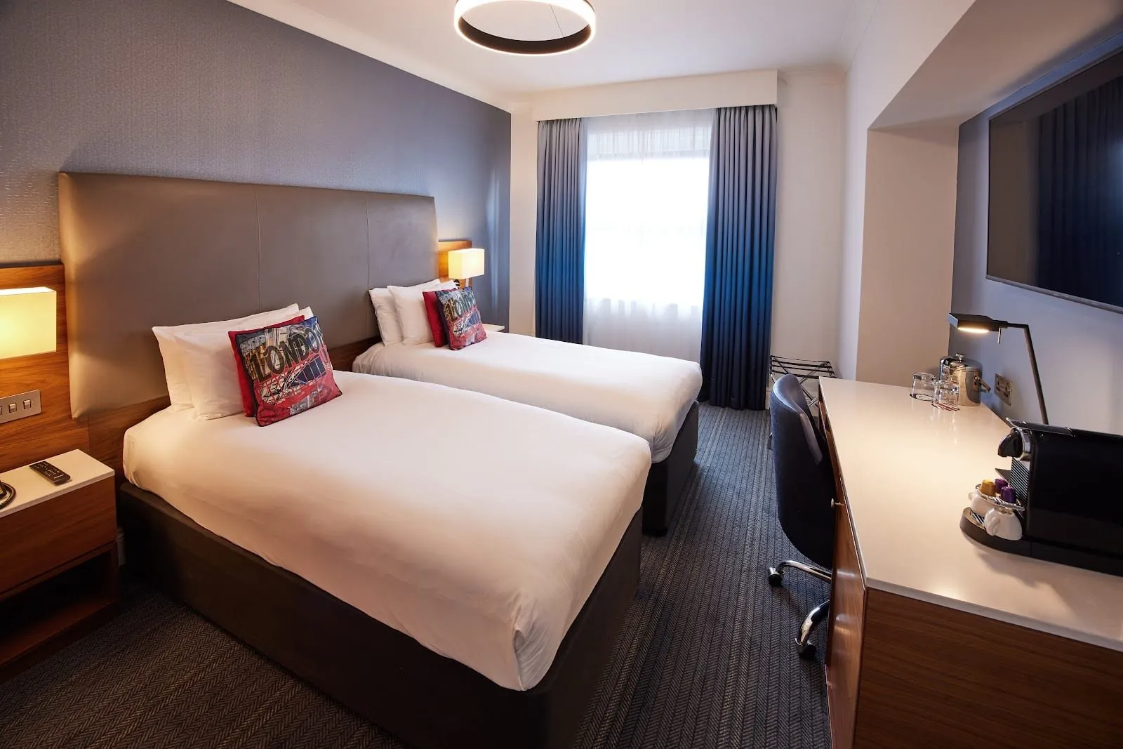 A twin room at The Cumberland with two single beds against a grey leather headboard, red London-print cushions, blue curtains at the far window and a long desk with a coffee machine under a wall-mounted TV