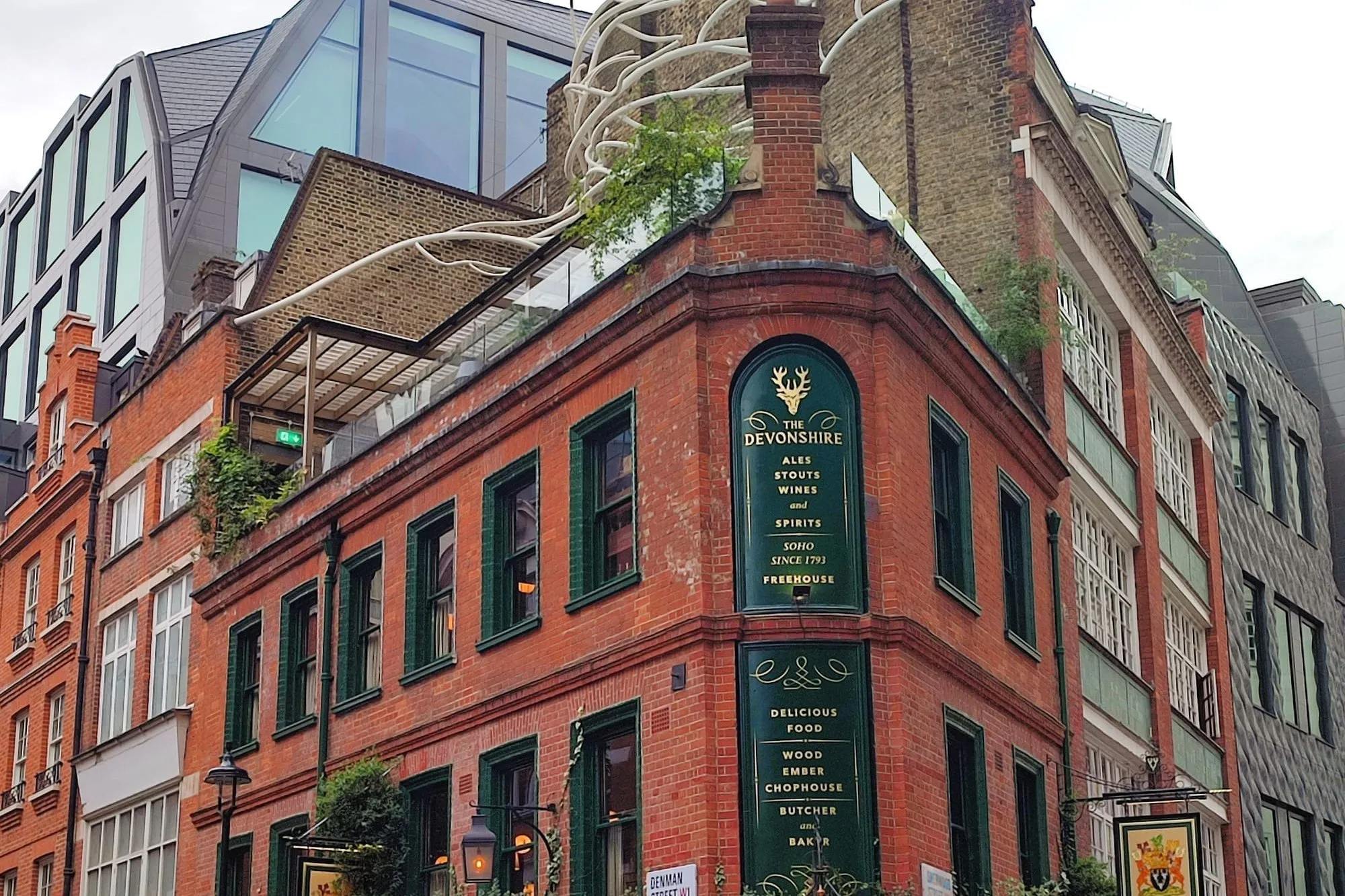 The Devonshire on the corner of Denman Street in Soho: a red-brick Victorian pub with dark green tiling and gold lettering reading 'Ales, stouts, wines and spirits, Soho since 1793', ivy along the ground floor and a planted roof terrace above, with a modern glass office building rising behind it