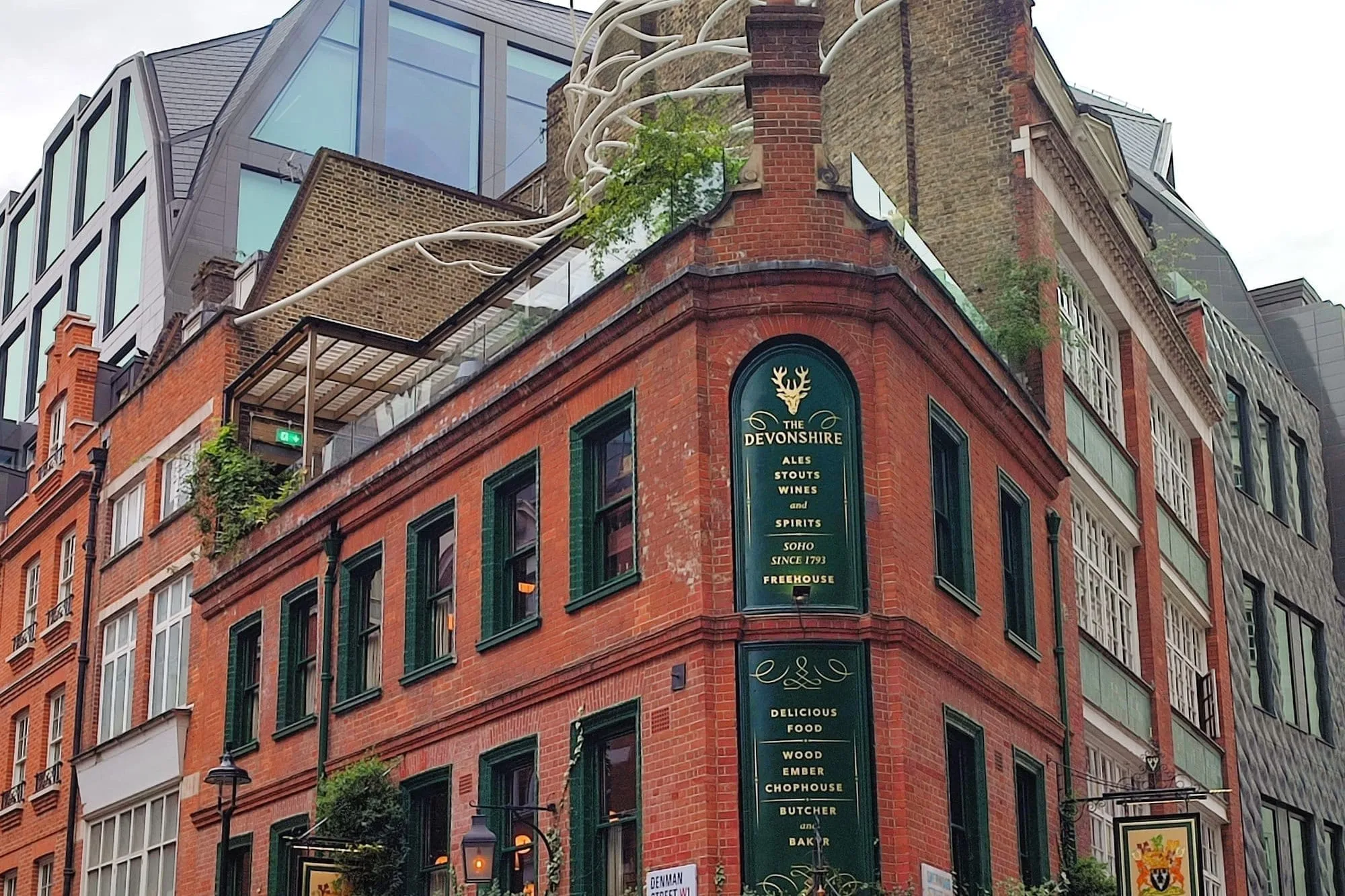 The Devonshire on the corner of Denman Street in Soho: a red-brick Victorian pub with dark green tiling and gold lettering reading 'Ales, stouts, wines and spirits, Soho since 1793', ivy along the ground floor and a planted roof terrace above, with a modern glass office building rising behind it
