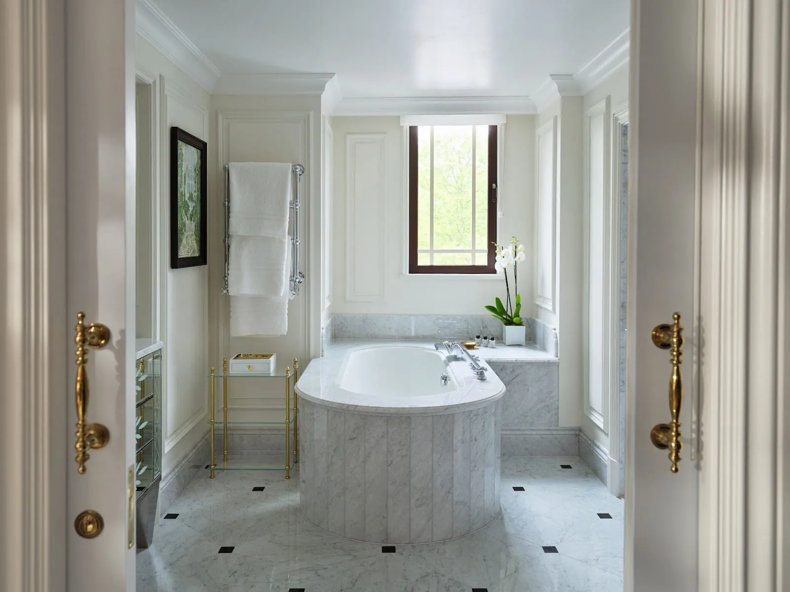 A Dorchester bathroom in white Carrara marble with an oval bath in a marble surround beneath a window onto trees, a heated towel rail and a floor inset with black squares