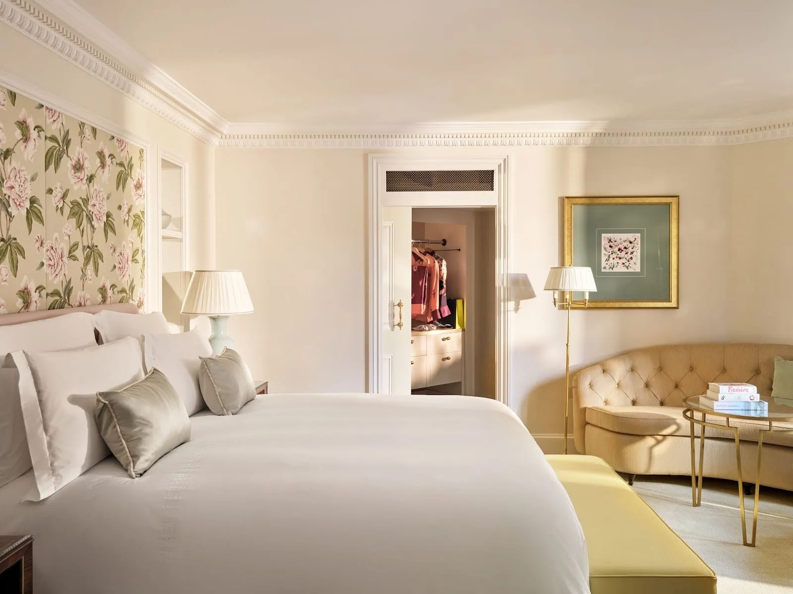 A Dorchester bedroom with a peony-print fabric panel behind a wide white bed, a buttoned cream sofa, a brass and glass side table and a door open to a walk-in wardrobe