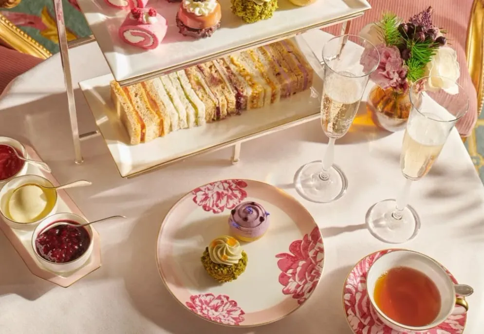 A tiered stand of finger sandwiches and pastries on floral china, with champagne flutes and a pink and gold table setting