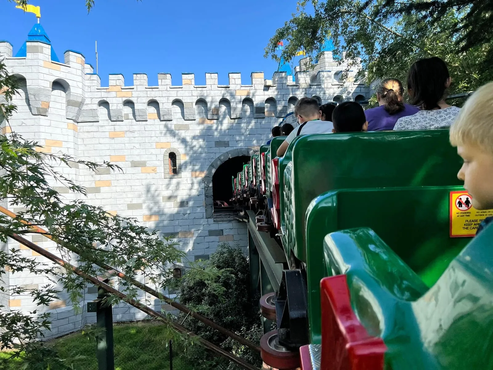 Riders in a green dragon-fronted coaster train passing the grey LEGO-style castle walls of Knights' Kingdom
