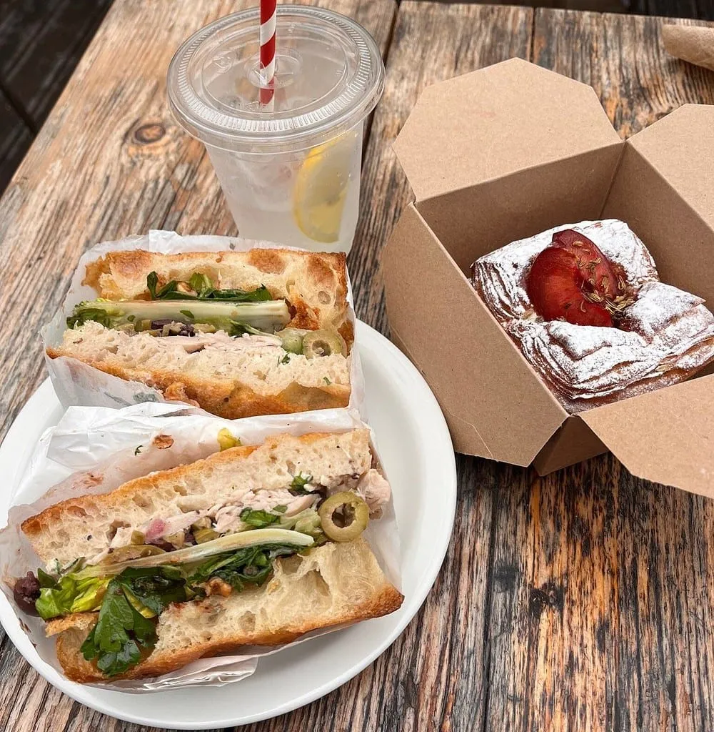 A focaccia sandwich cut in half on a white plate, open-crumb bread around chicken, green olives, herbs and leaves, with a lemonade and a plum pastry in a box alongside