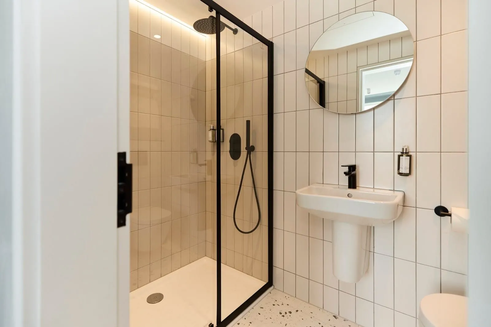 A shower room at The Edwin with white vertical tiles, a walk-in shower with a black rain head behind a black-framed glass door, and a round mirror above a small basin