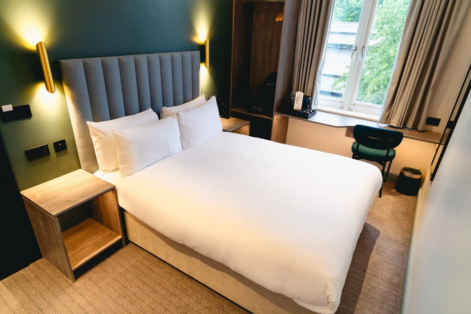 A double room at The Edwin with a grey channelled headboard, brass wall lights on a dark green wall, open wardrobe shelving and a desk under a window facing trees