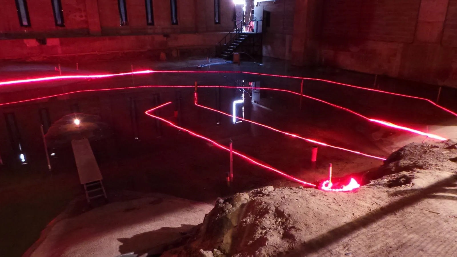 The flooded remains of the Rose in a dark basement, red rope lights tracing the lines of the playhouse walls and stage across the water, with mounds of earth in the foreground