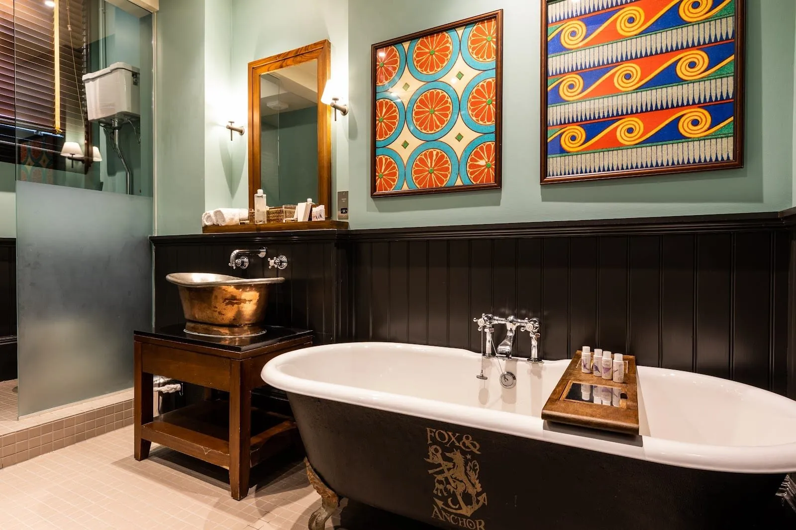 A bathroom at The Fox and Anchor with a black roll-top bath bearing the pub's crest, a hammered copper basin on a wooden stand, black panelling, mint walls and two bold framed prints
