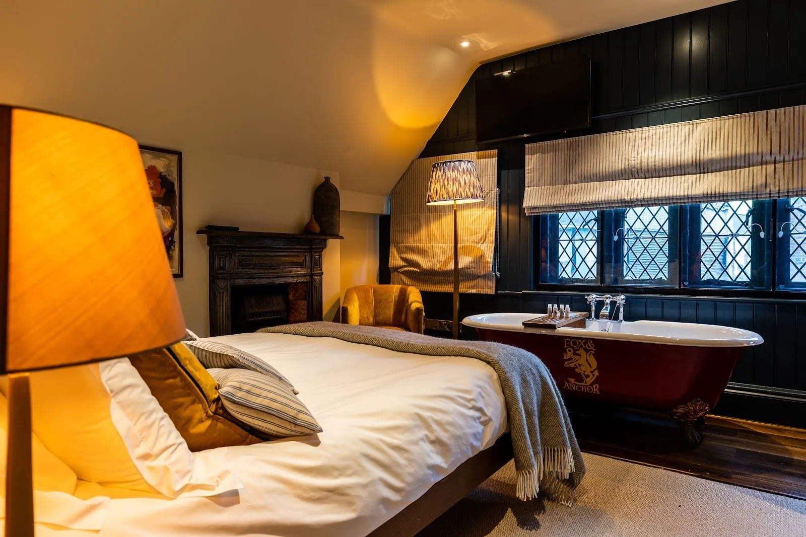 A bedroom at The Fox and Anchor with a red roll-top bath bearing the pub's crest beside the bed, dark green panelling, leaded diamond-pane windows, a cast-iron fireplace and a mustard velvet armchair