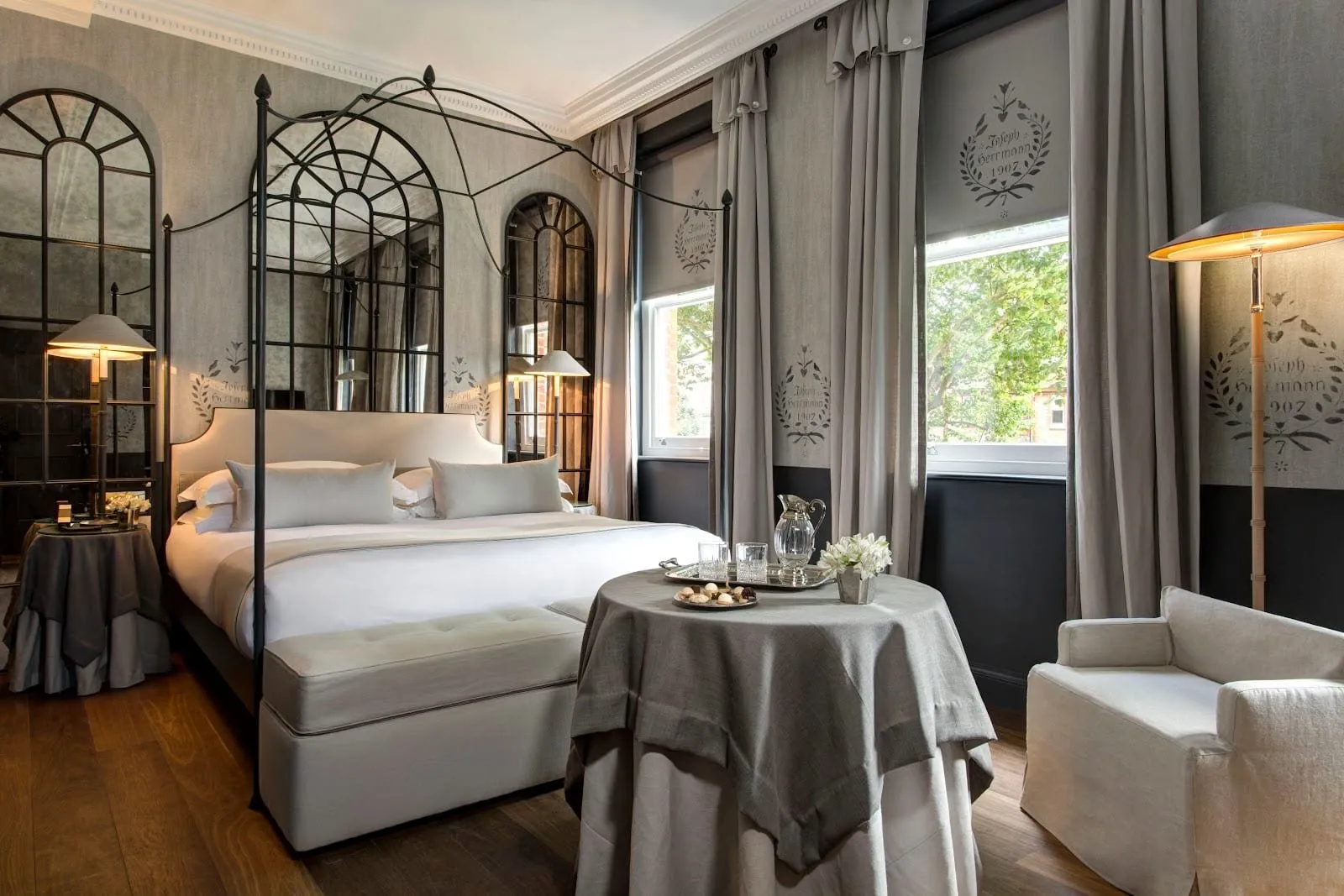 A Superior room at The Franklin: a black iron four-poster bed, arched antiqued mirrors behind it, grey linen walls stencilled with laurel wreaths, a round table with a grey cloth and two sash windows onto trees