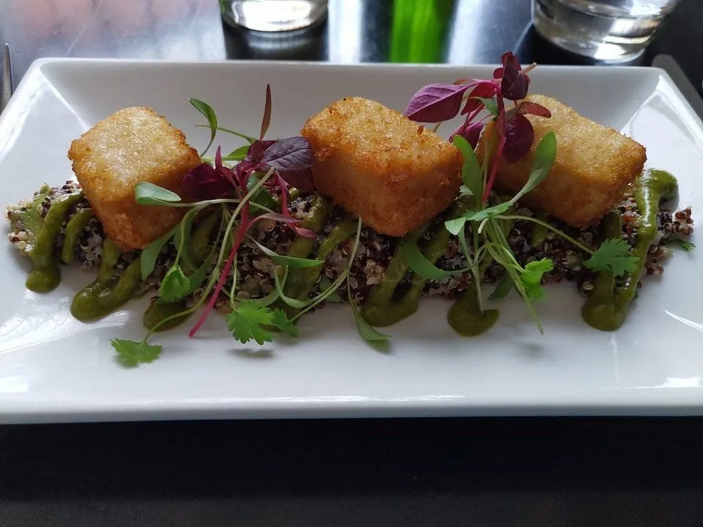 Three crisp-fried golden cubes set along a long white plate on quinoa, with a green herb sauce and micro leaves