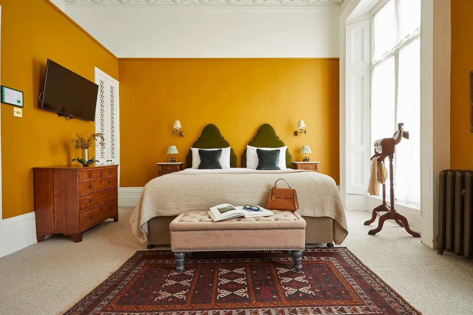 A high-ceilinged room at The Georgian House with mustard-yellow walls, a double bed with twin arched green velvet headboards, an antique chest of drawers, a red patterned rug and a tall shuttered sash window