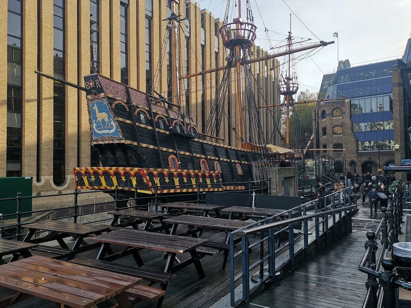 The Golden Hinde in St Mary Overie Dock, its black hull striped red and yellow, a golden deer on a blue shield on the stern, and three masts against an office block
