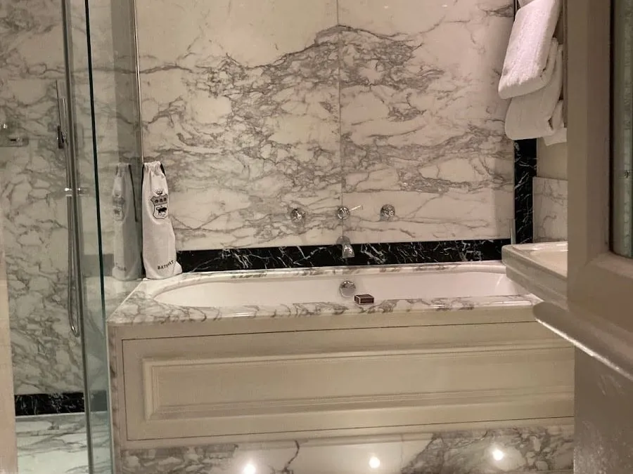 A bathroom at The Goring clad in white and grey veined marble, with a panelled bath and a separate glass-walled shower