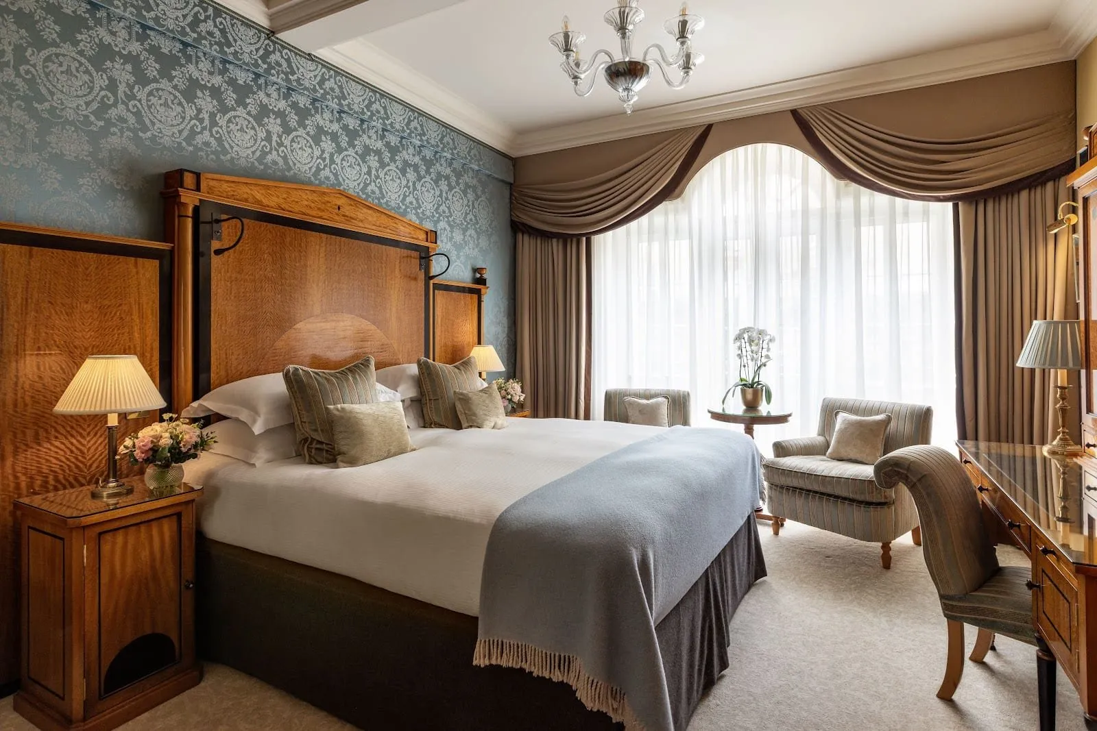 A room at The Goring with blue silk damask walls, a tall satinwood headboard, a pale blue throw, and two striped armchairs in a wide window hung with swagged curtains