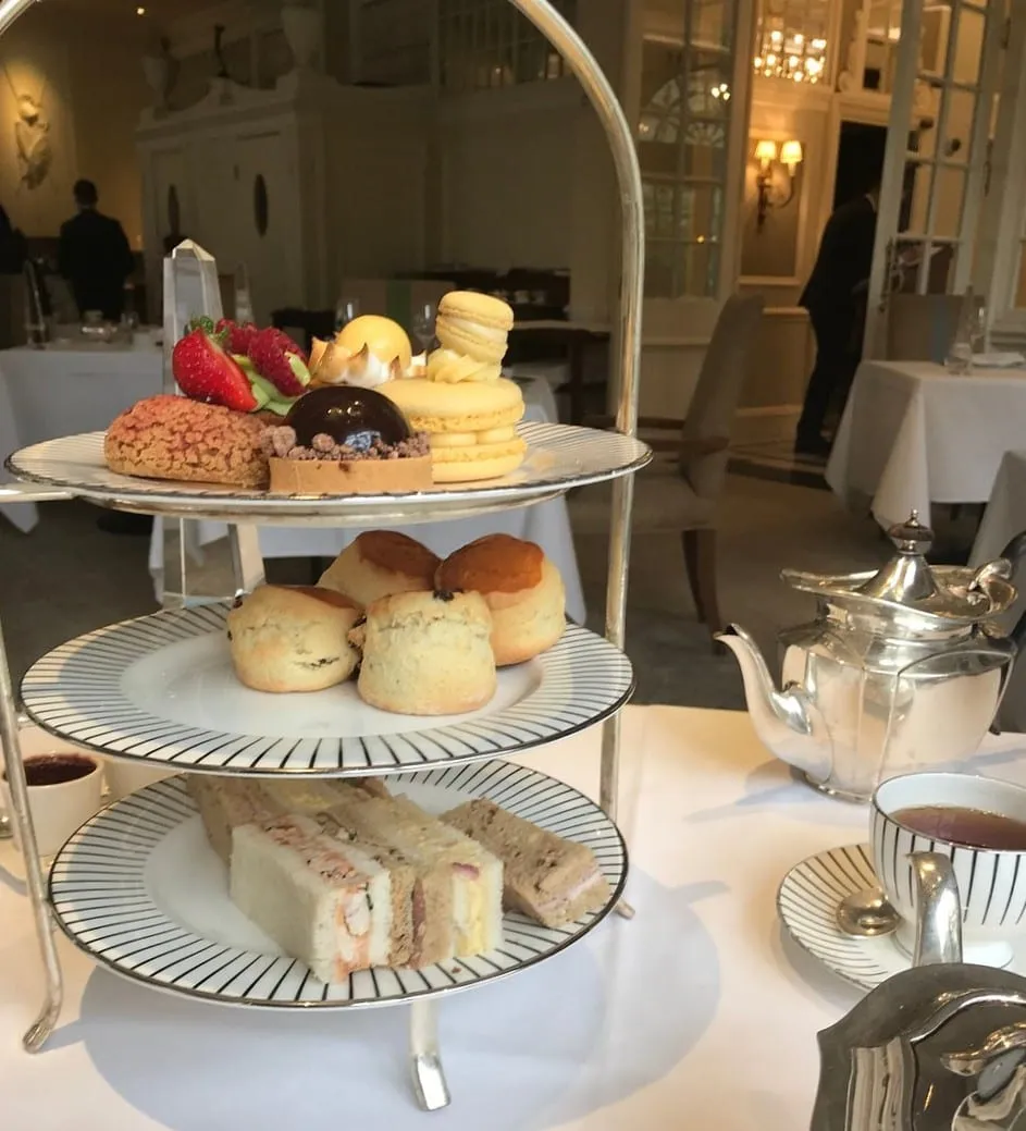 A tiered stand of finger sandwiches, scones and pastries on striped china in a bright white dining room, with a silver teapot alongside