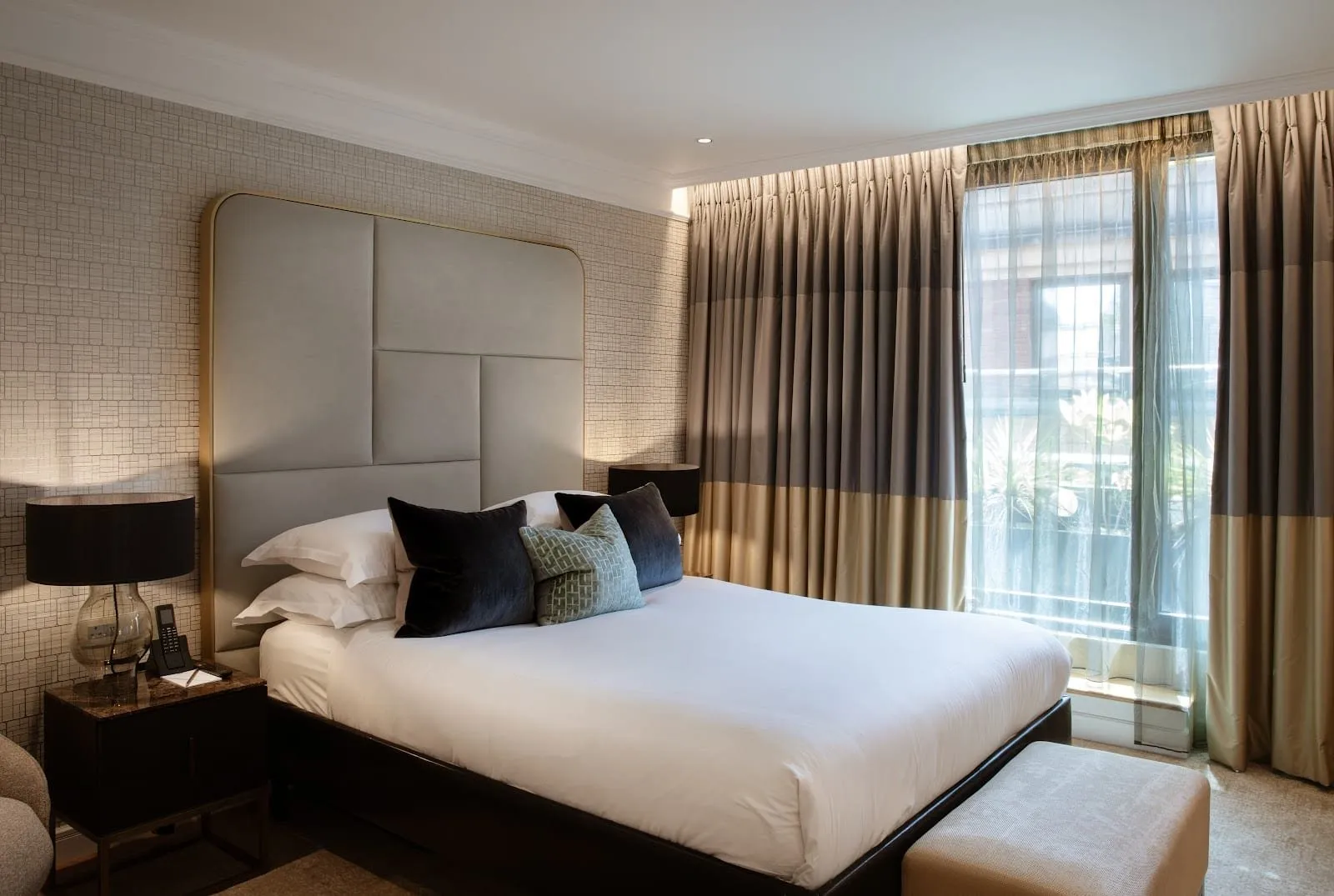 A double room at The Guardsman with a tall grey leather-panelled headboard edged in brass, dark velvet cushions, grid-patterned wallpaper and a full-height window with sheer and striped curtains
